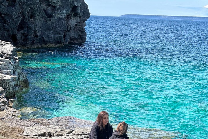 a group of people sitting on a rock near the ocean with Bruce Peninsula National Park in the background