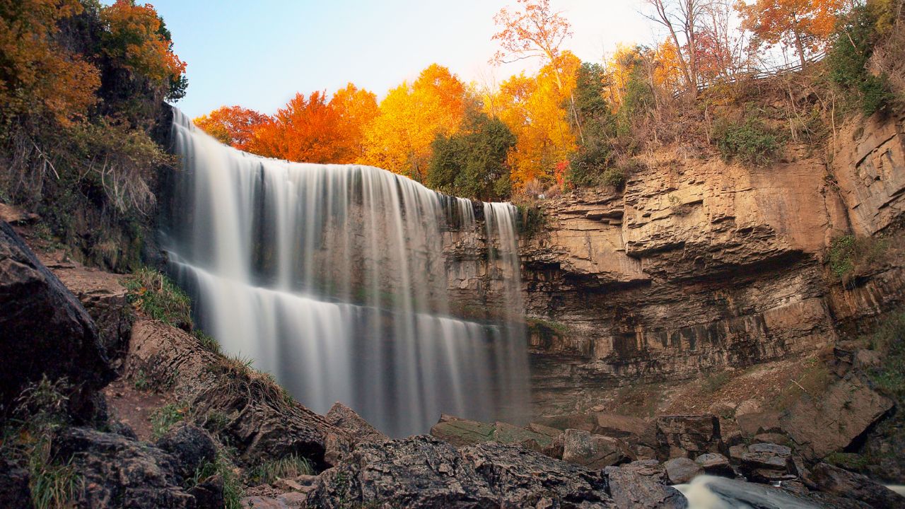 Waterfall flowing down rocky cliff with autumn trees in background.