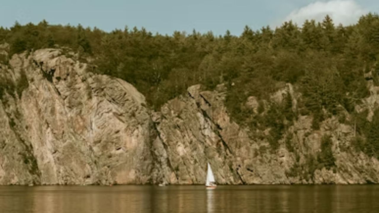 Sailboat on calm water near rocky cliff with trees under a clear sky.