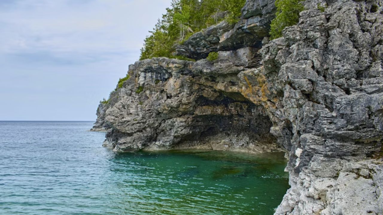 Rocky cliff with green water and trees under an overcast sky.