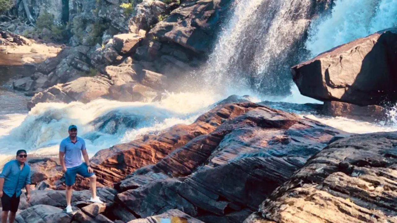 Two people on rocky terrain near a cascading waterfall on a sunny day.
