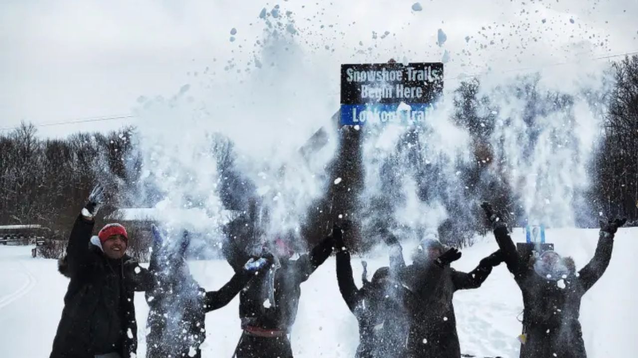 People in winter clothing throw snow in the air, standing in front of a sign for snowshoe trails.