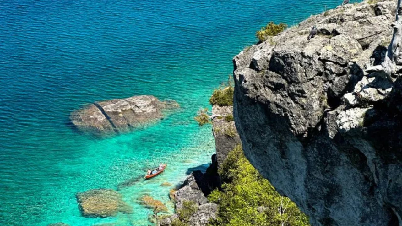 Kayaker paddling in clear turquoise water below a high rocky cliff
