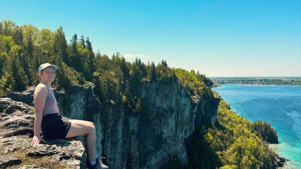 Woman sitting on cliff edge overlooking forested coastline and blue water at Lion’s Lookout