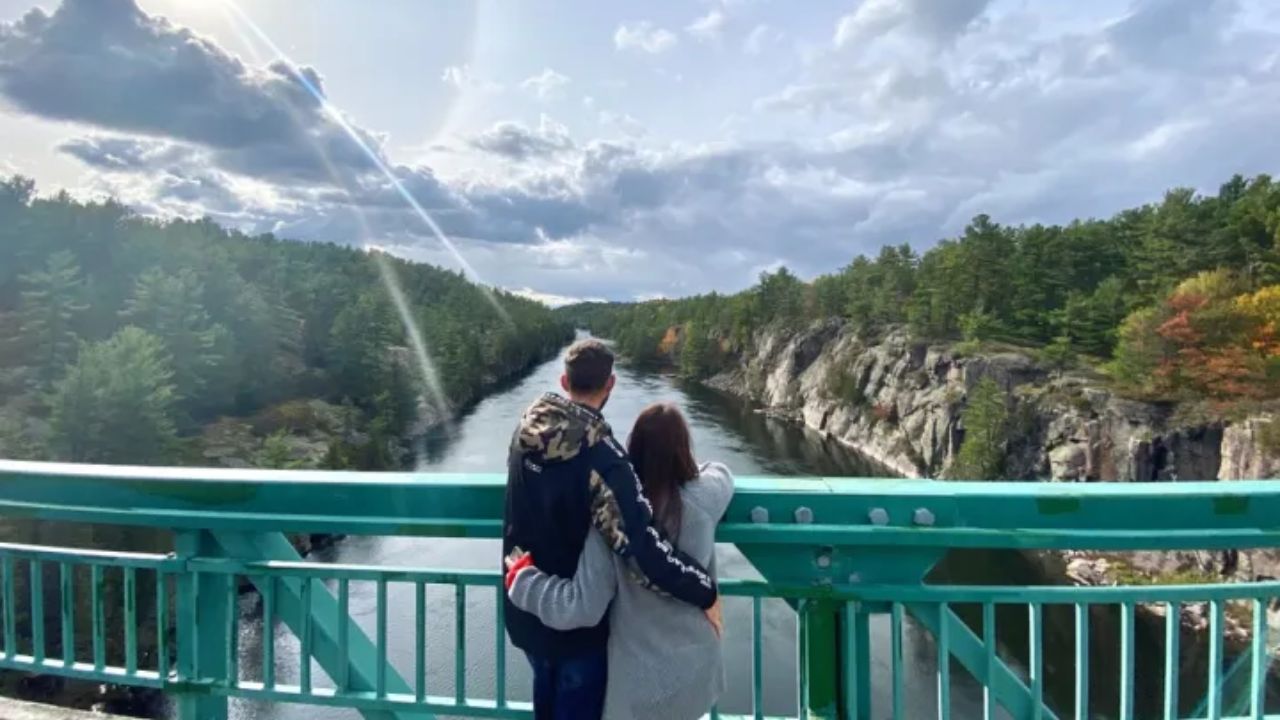 A couple standing on a green bridge looking over the French River flowing between rocky cliffs and forest
