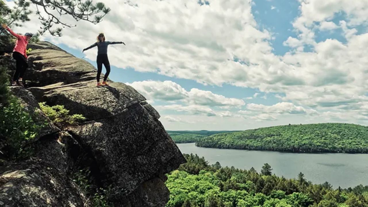 Hikers standing on rocky cliffs with scenic lake and forest view below.
