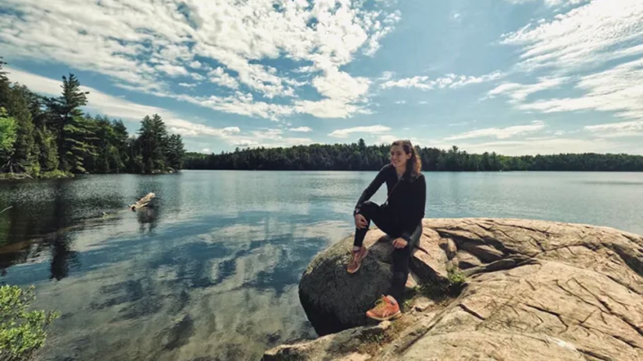 Woman sitting on lakeside rock with calm water and cloudy sky reflection.
