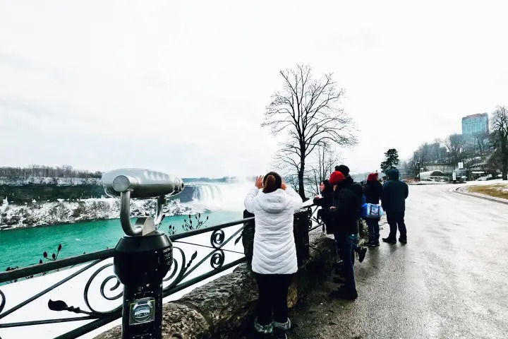 Visitors standing at a Niagara Falls lookout point in winter, taking photos near the railing overlooking the falls