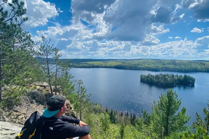 Backpacker resting at a high-elevation viewpoint at Centennial Ridges Trail above a lake surrounded by dense northern forest.