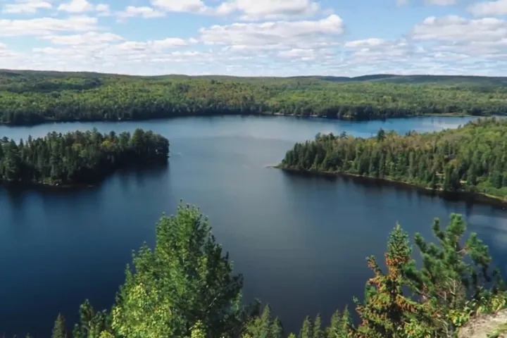 Scenic aerial view of a forest-lined lake from a rocky overlook in Algonquin Provincial Park under a partly cloudy sky.