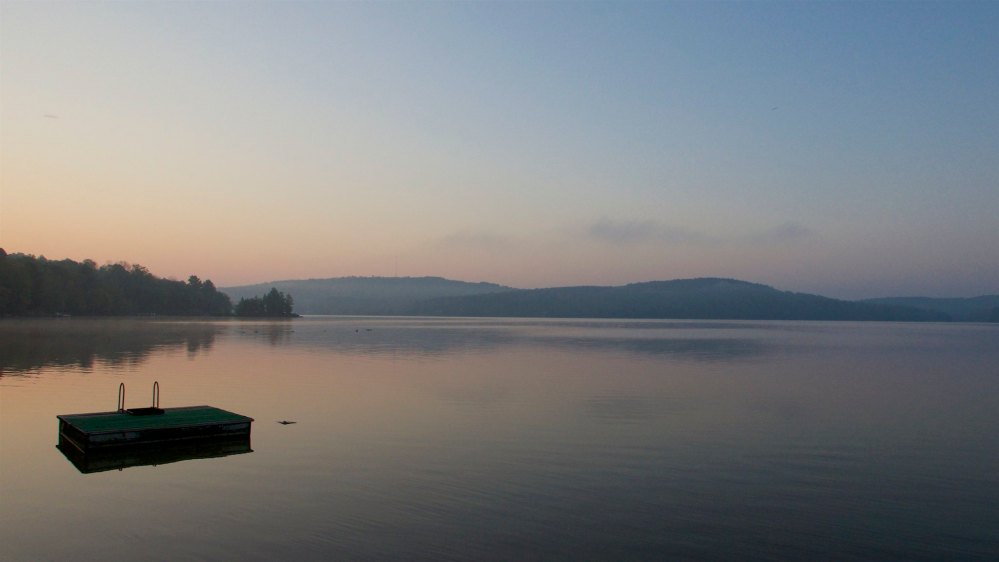Calm Ontario lake at dusk with a floating dock, distant forested hills, and soft pastel skies.