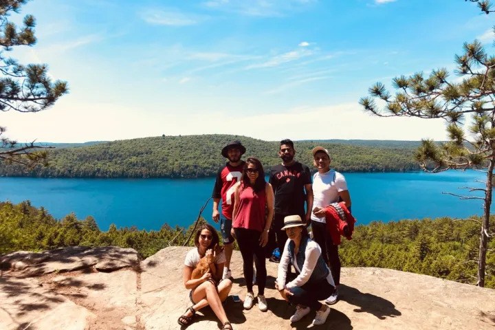 Group of hikers posing at a scenic rocky lookout overlooking a bright blue lake and forested landscape in Ontario.