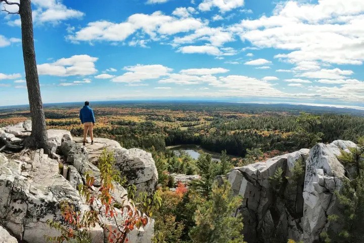 Hiker overlooking rugged quartzite cliffs, colourful forest, and distant lakes from a scenic Ontario hiking trail.