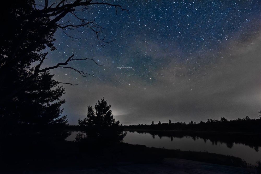 Star-filled night sky reflected over a peaceful lake surrounded by silhouetted trees in Ontario.