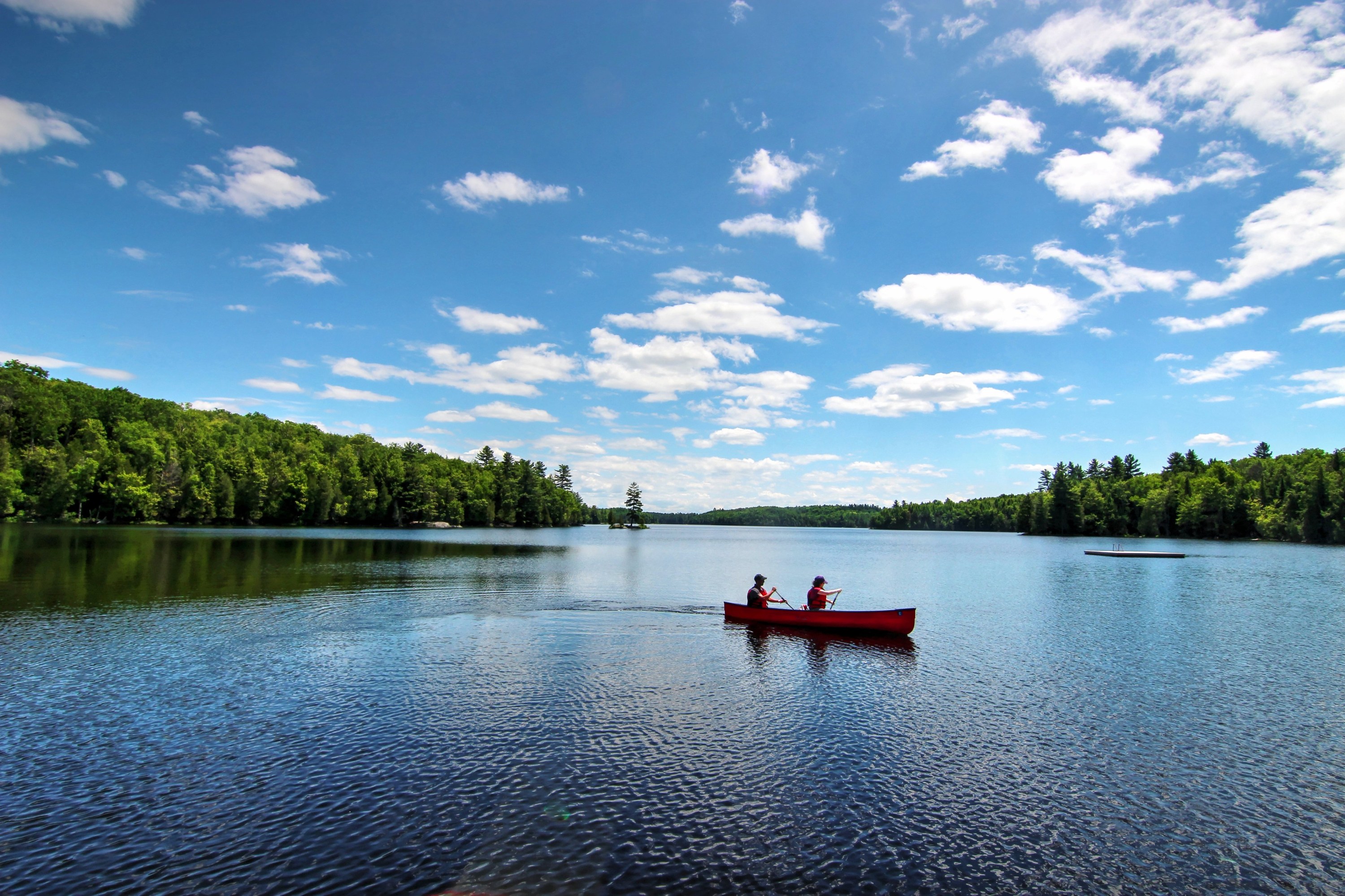 Two people paddling a red canoe in Silent Lake Provincial Park surrounded by lush green forest under a blue cloudy sky