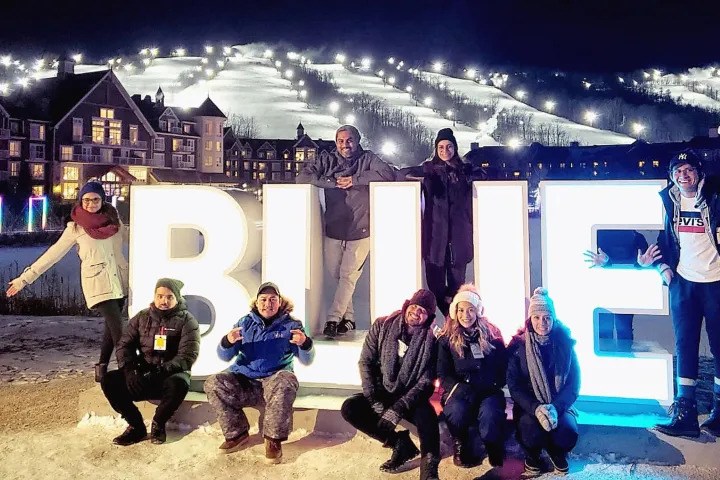 Group of friends in winter coats posing around large illuminated BLUE letters at Blue Mountain Resort at night.