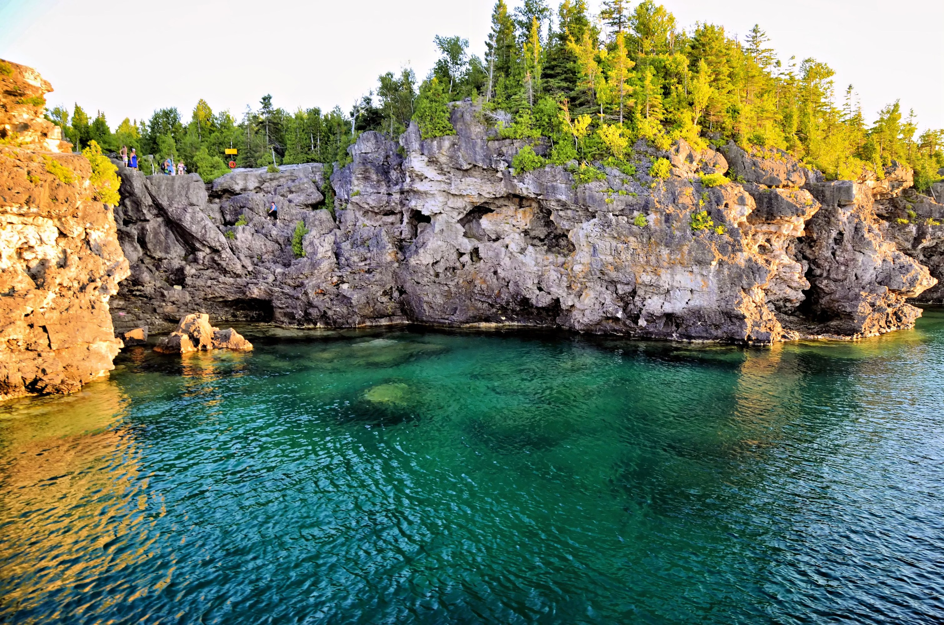Sunlit rocky cliffside cove with green trees along the edge overlooking clear emerald green water