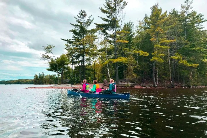 Three people paddling a canoe across a lake lined with pine trees at Bon Echo